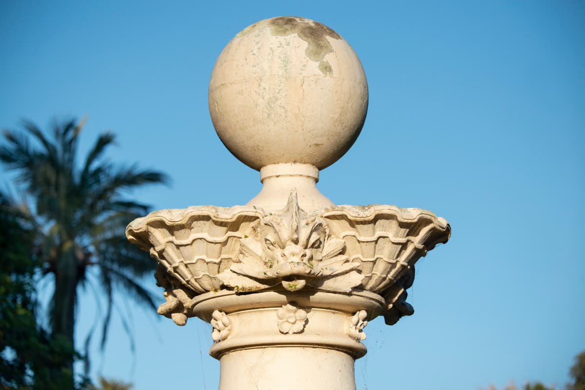 Close-up of stone fountain detail with carved column and palm trees behind.